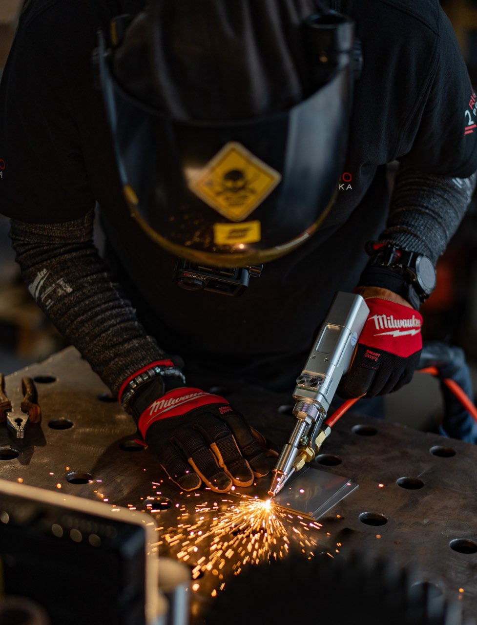 Spawanie i czyszczenie laserowe. Spawacz w przyłbicy i rękawicach Milwaukee wypalający metal, iskry rozbłyskują nad stołem warsztatowym